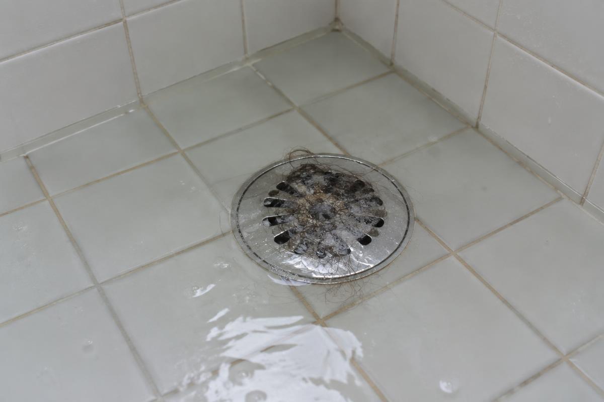 Standing water over a clogged chrome shower drain in a white tiled residential shower with visible hair buildup